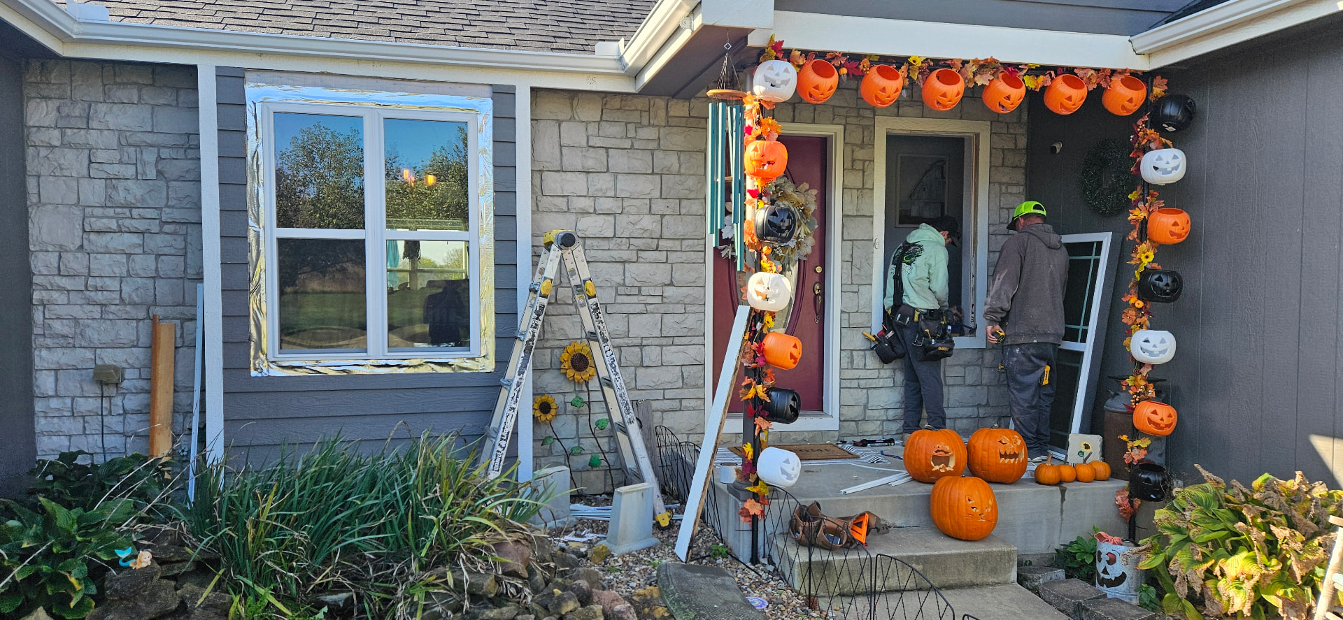 Two Kan Exteriors workers fitting a replacement window at a home with gray siding and a stone veneer front, silver flashing tape around the new opening and an aluminum ladder in the yard