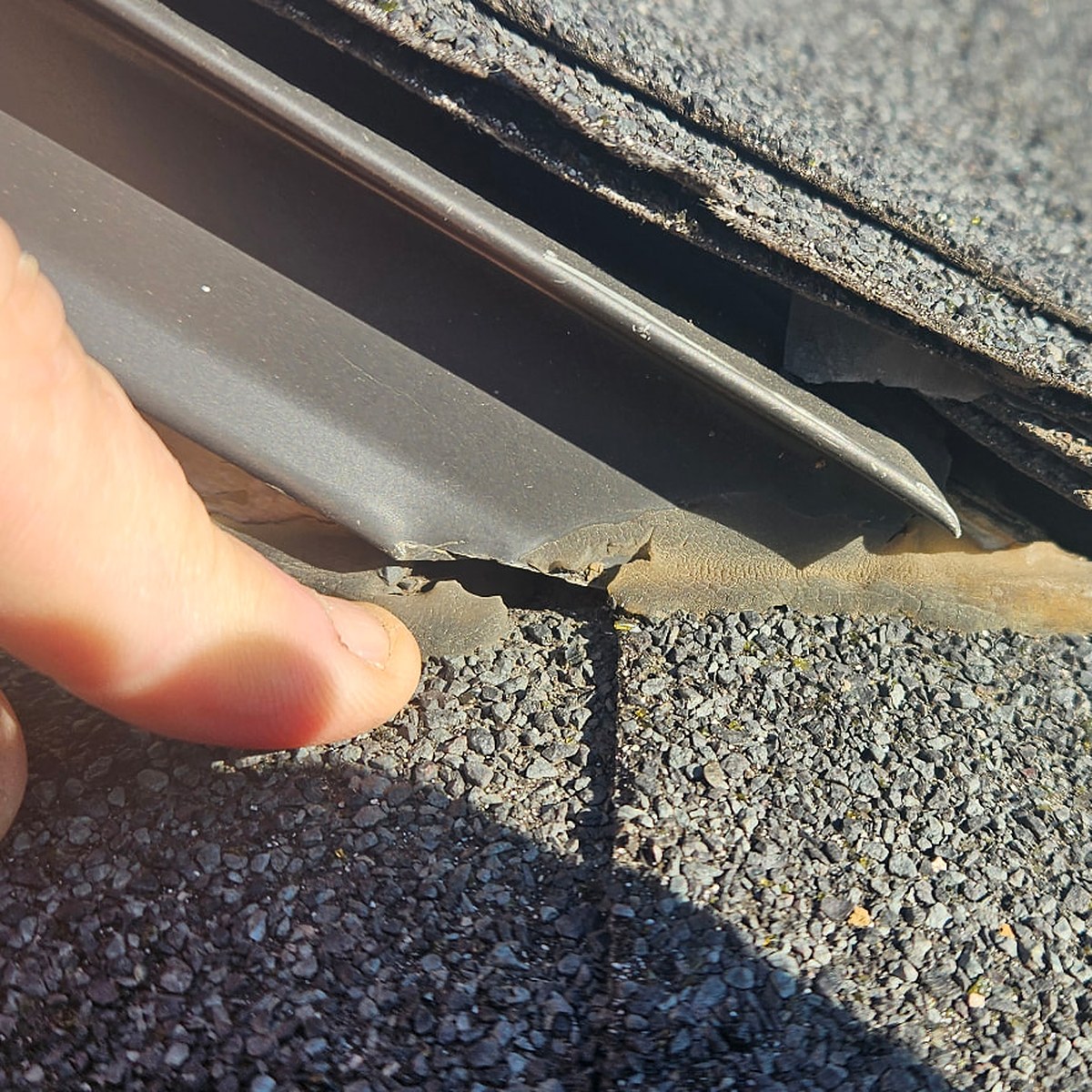 Homeowner's hand pointing at a lifted, cracked flashing edge on an asphalt shingle roof